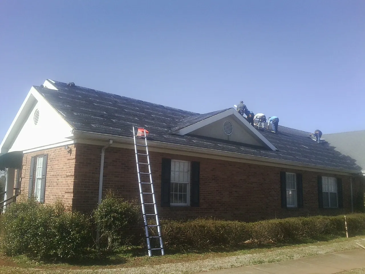 Crew tearing shingles off a brick ranch roof with a ladder set — Emergency Roof Repair work by our crew in Center Barnstead