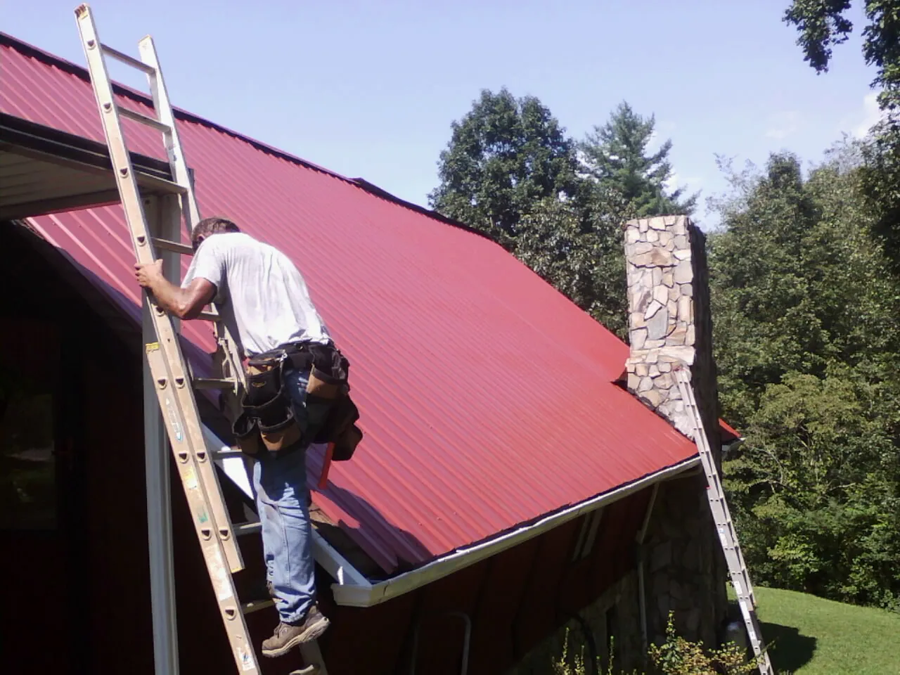 Roofer on a ladder beside a red metal roof and stone chimney — Soffit & Fascia Repair by Roofing Brothers in Center Barnstead
